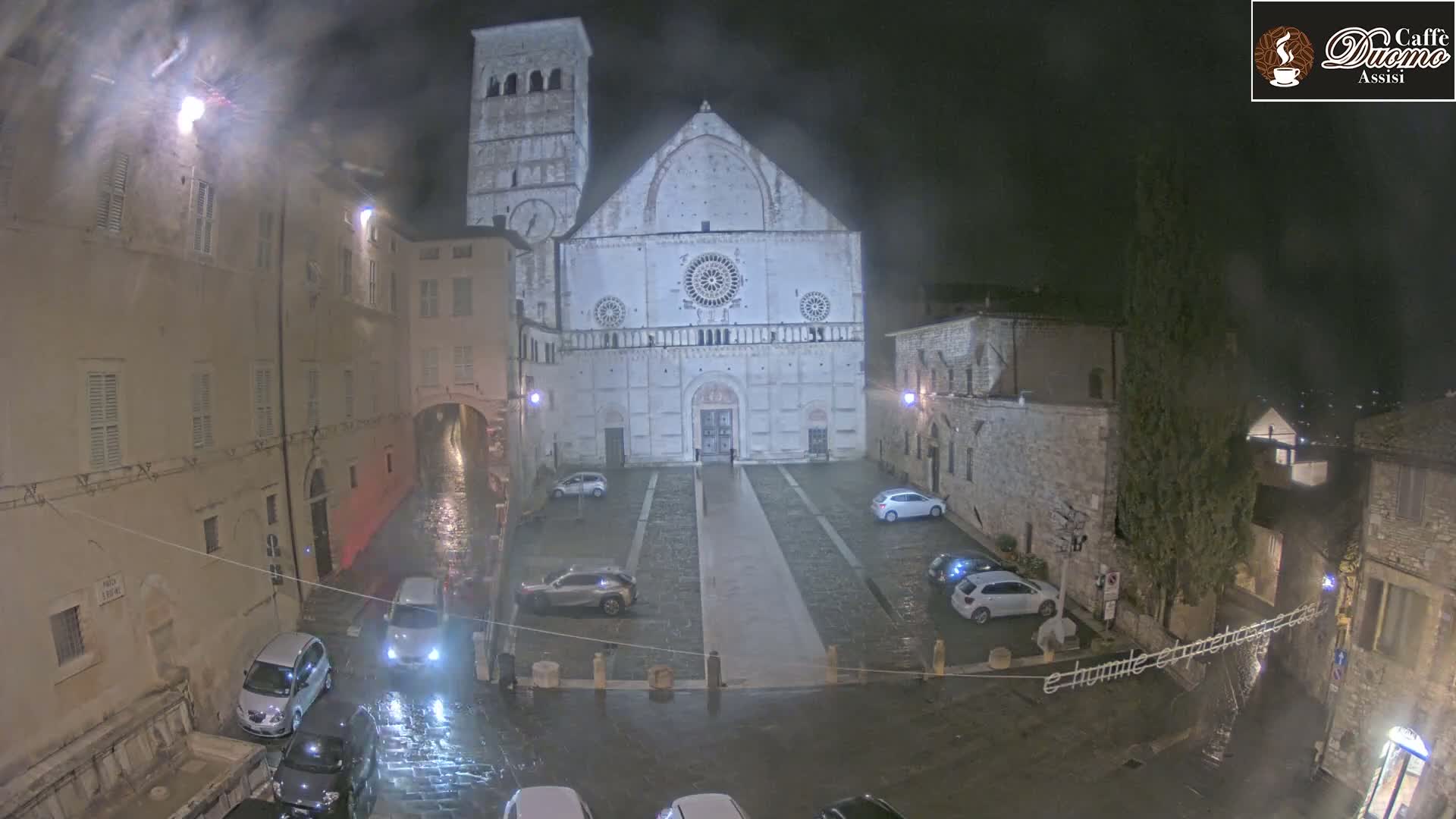 A large stone cathedral stands in a rain-soaked town square at night, surrounded by parked cars and older buildings.
