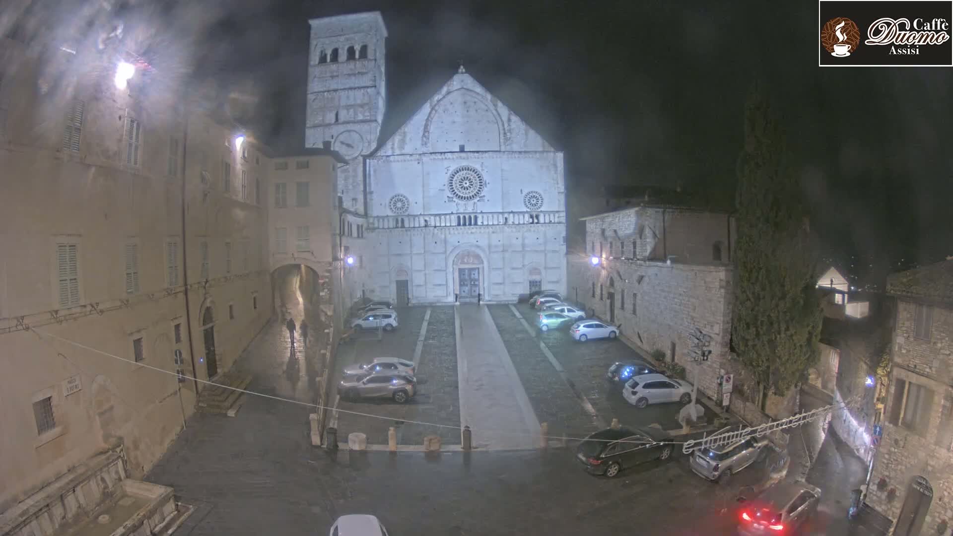 A large stone cathedral is visible at night in a town square with parked cars under a misty, rainy sky.