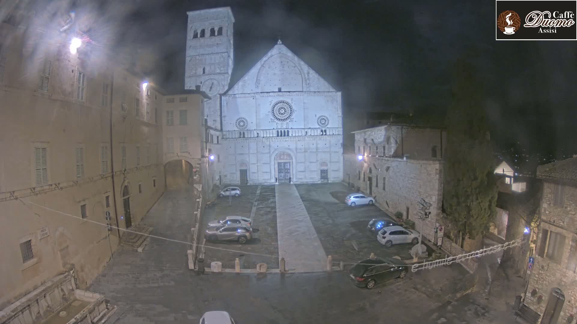 A large, light-colored cathedral stands in a dimly lit, empty square at night, with several cars parked around it and older stone buildings surrounding the square under overcast conditions.