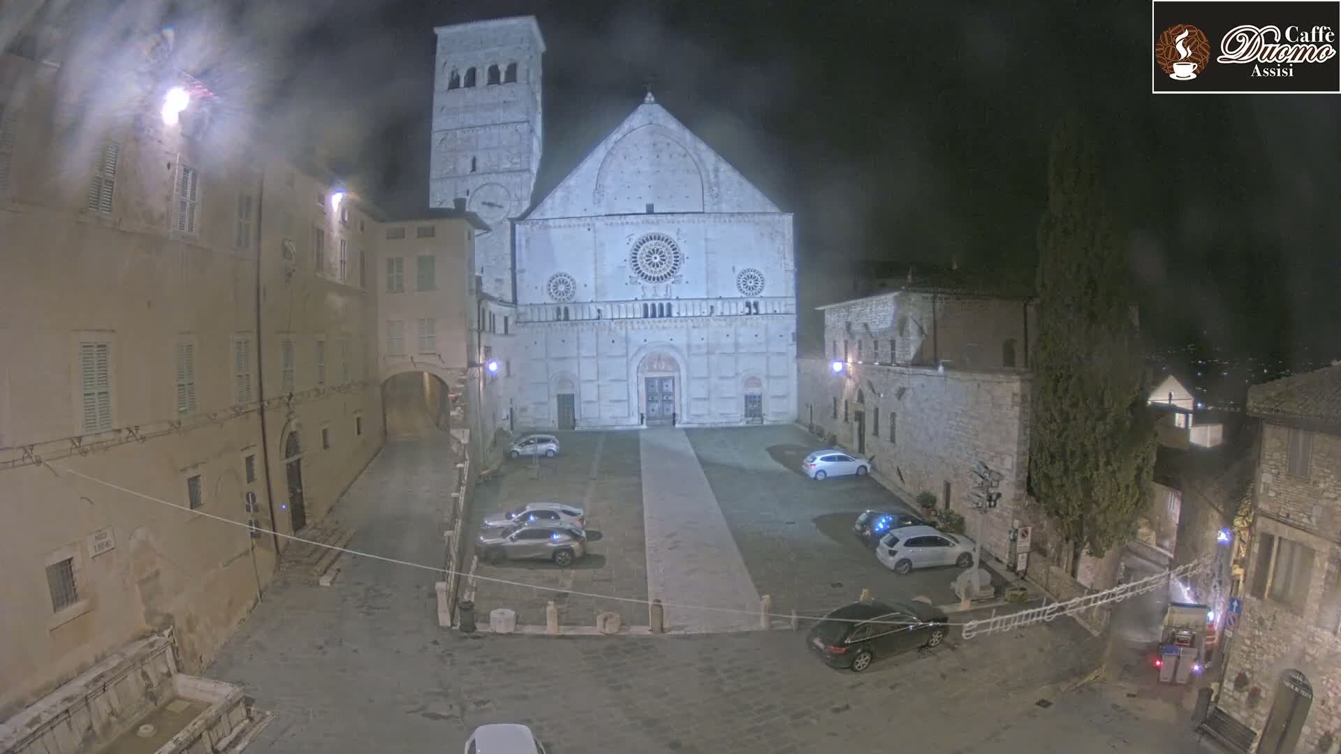 A large stone cathedral is illuminated at night in a town square with several cars parked, under overcast skies.