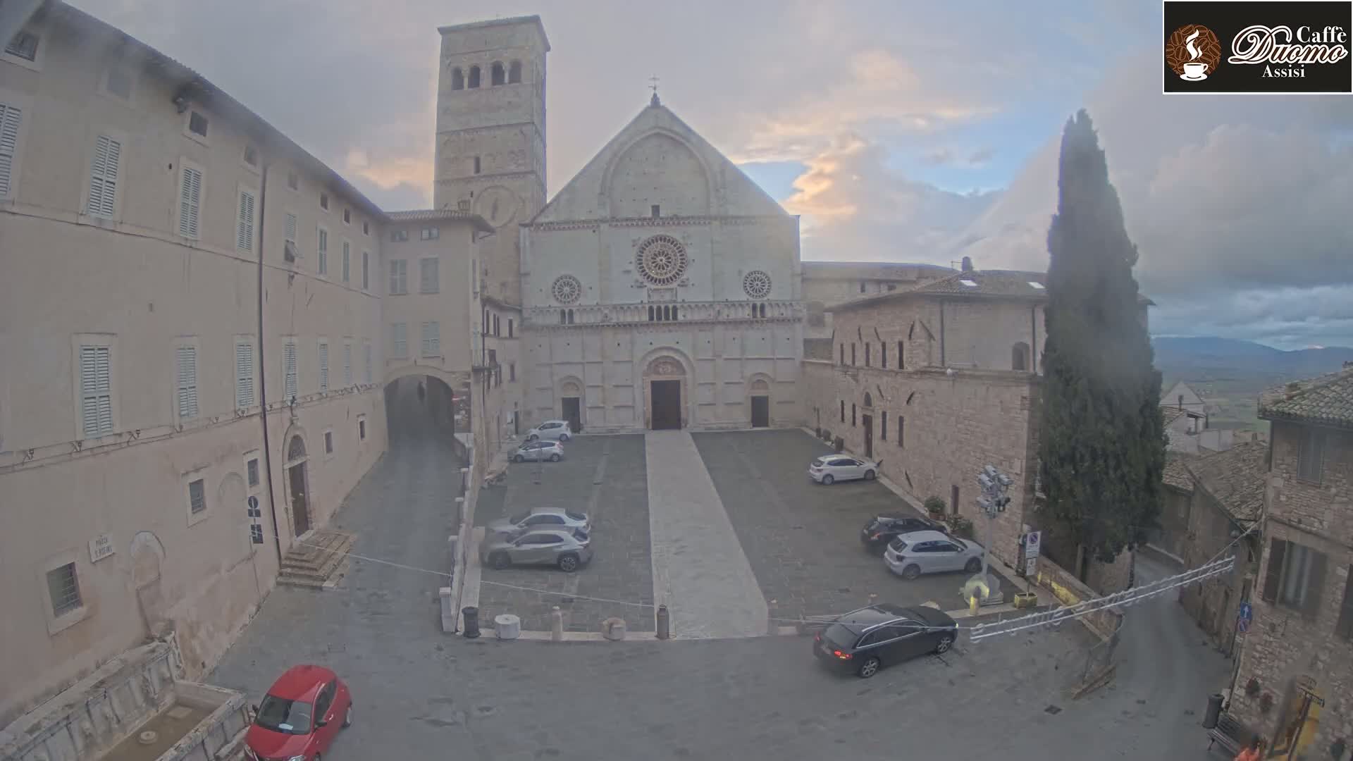 A large, light-colored cathedral stands in a courtyard surrounded by stone buildings under a cloudy sky.