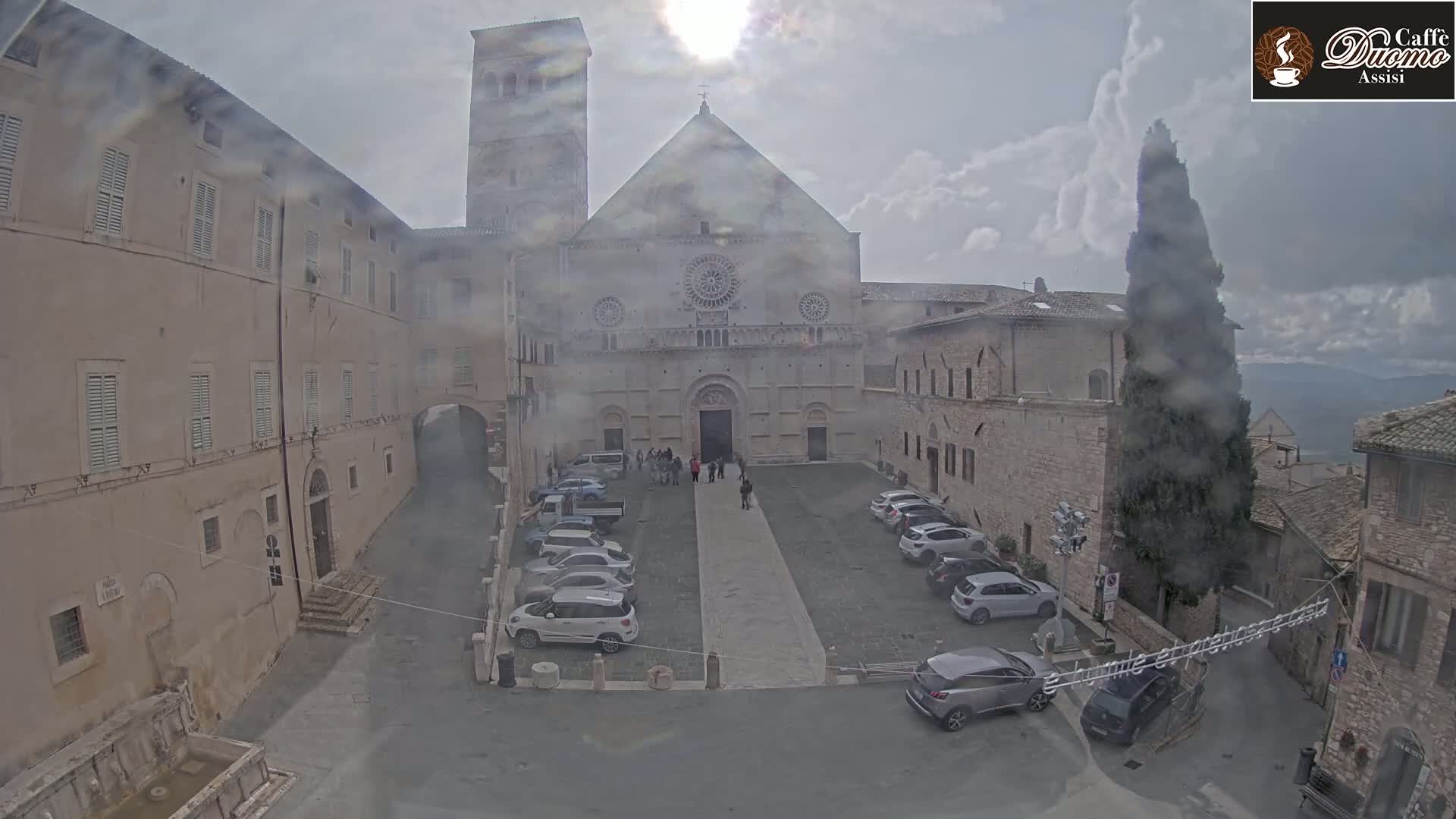 A large stone cathedral is situated in a town square with several cars parked, under an overcast sky.