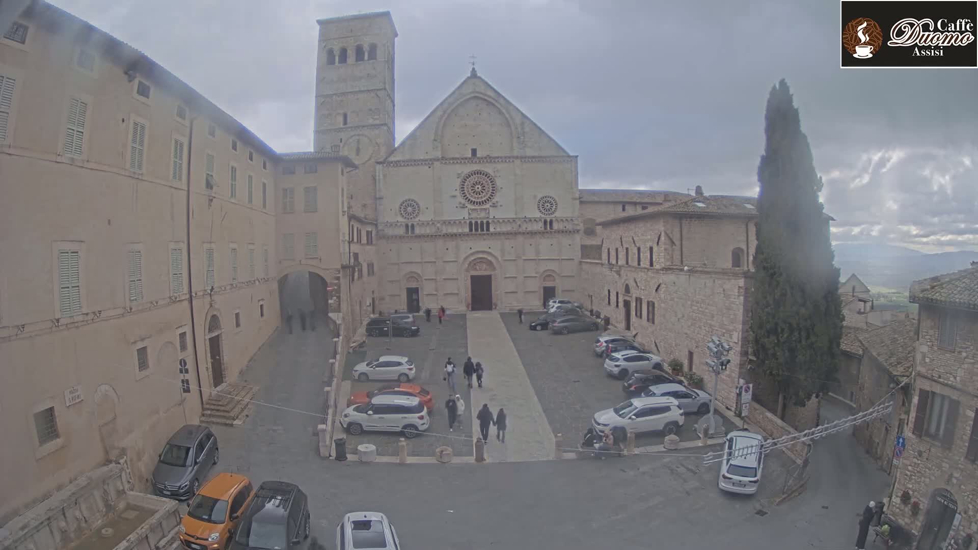 A large stone cathedral with a bell tower stands in a town square surrounded by old buildings on an overcast day.