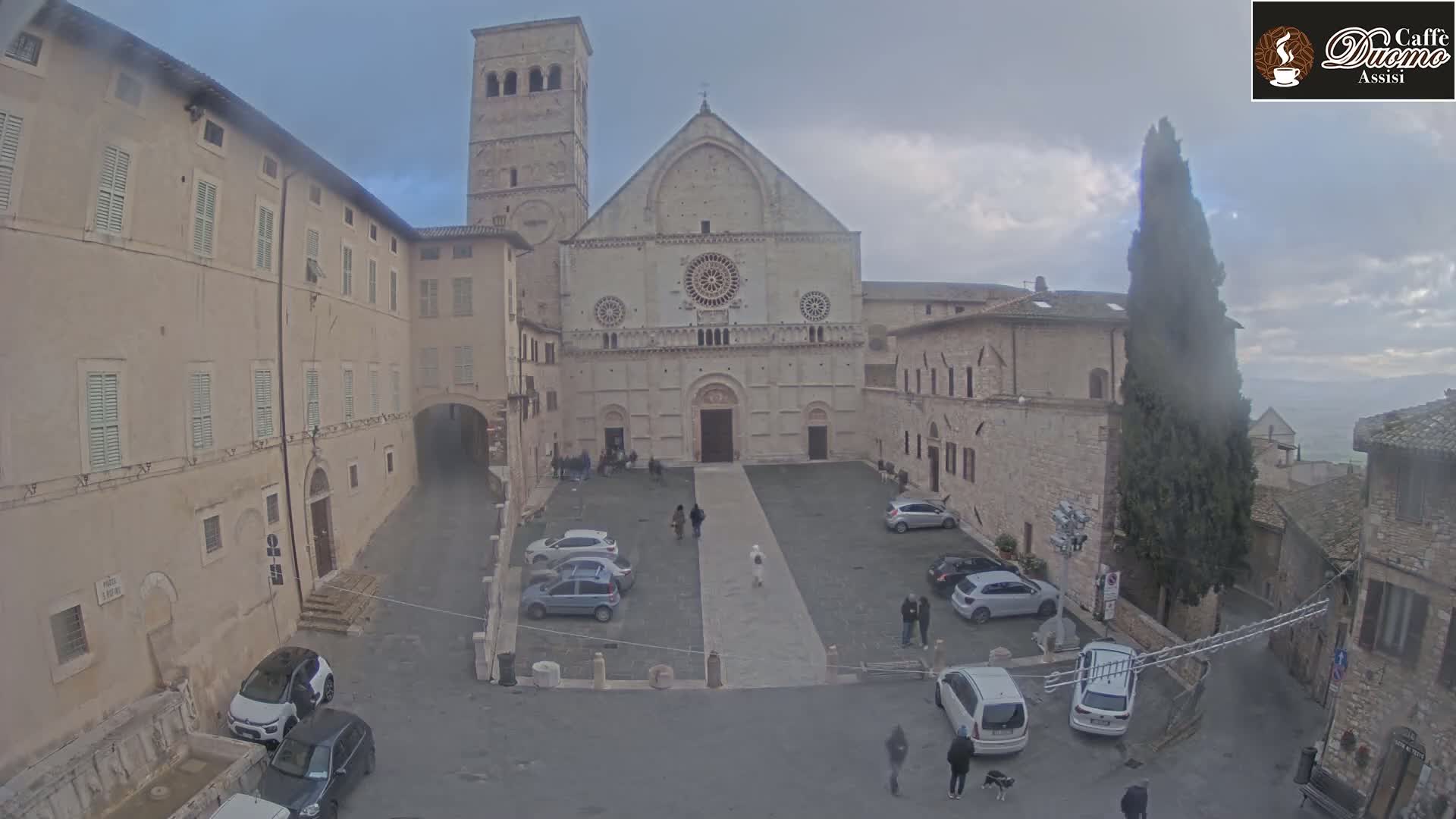 A large stone cathedral sits in a square surrounded by buildings under an overcast sky.