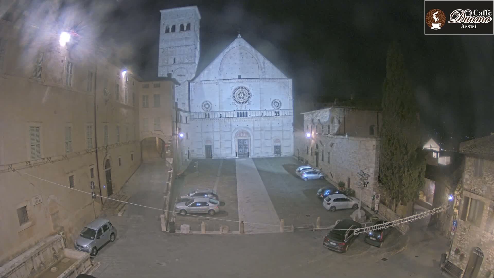 A large stone cathedral is situated in a town square at night, under a misty or hazy sky, with several cars parked around the square.