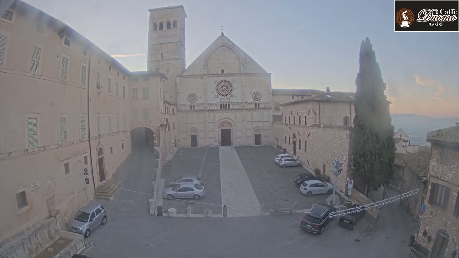 A hazy, pale-light outdoor view shows a large stone cathedral in a town square with several cars parked around it and buildings lining the square.