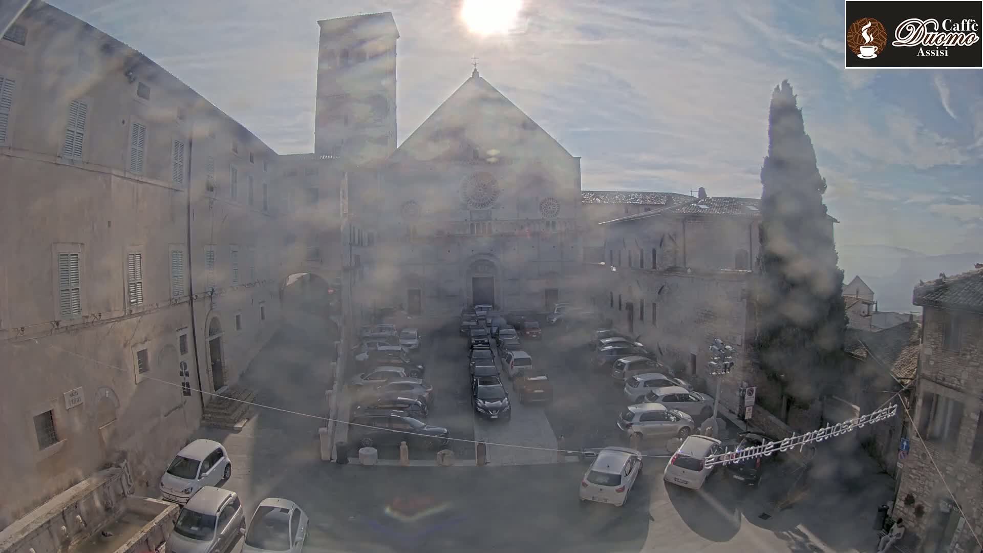 A hazy sun shines on a courtyard filled with parked cars in front of a large stone church and surrounding buildings.
