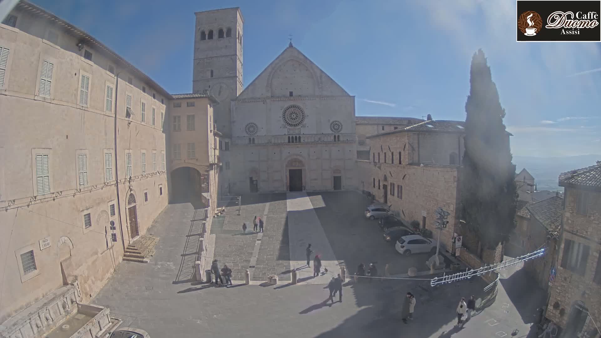 A large, light-colored stone church with a tall bell tower stands in a sunny town square, surrounded by old buildings and a few pedestrians.