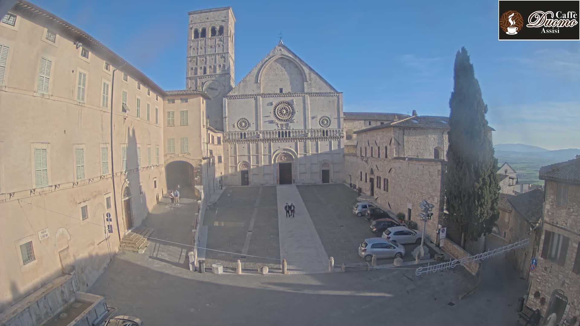 A large stone cathedral stands in a sunlit town square, surrounded by buildings and a few parked cars under a clear blue sky.