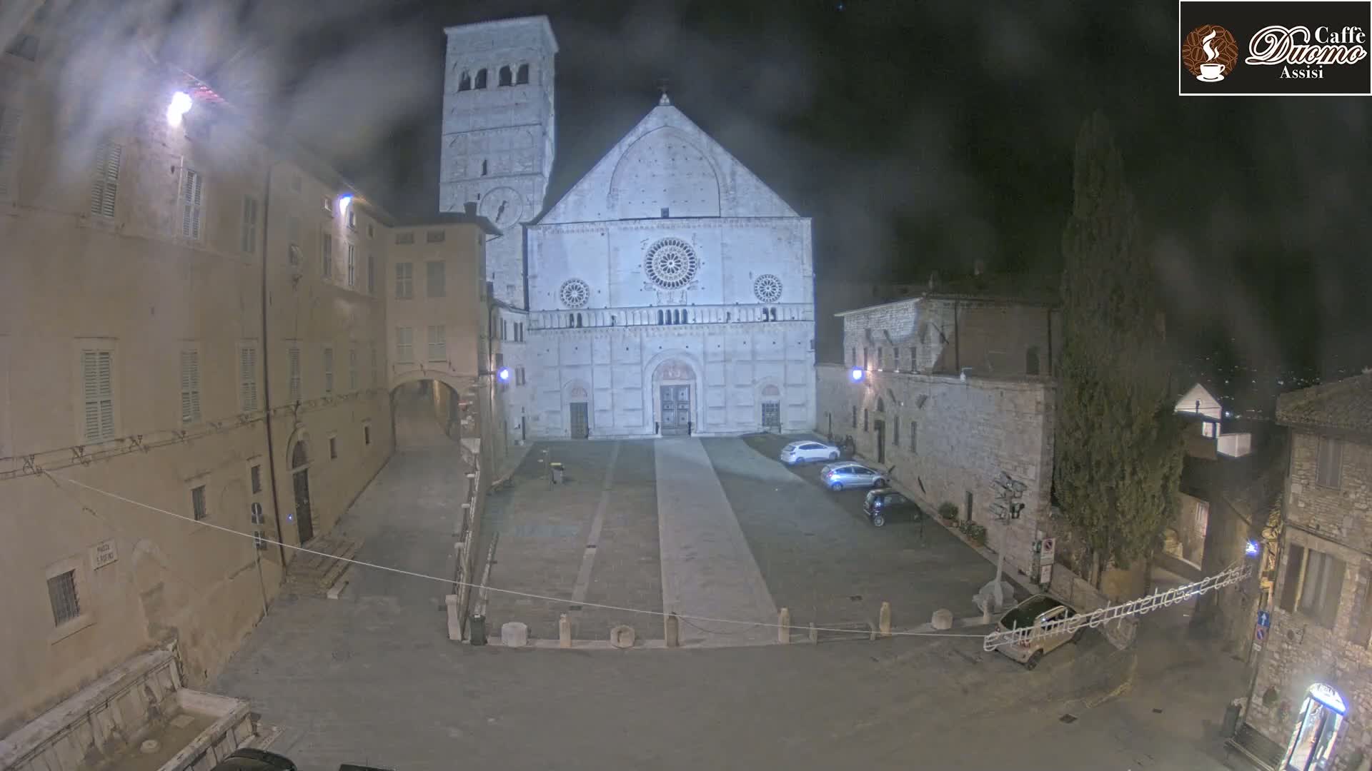 A large stone cathedral is seen at night in a mostly empty square, under hazy or foggy conditions.