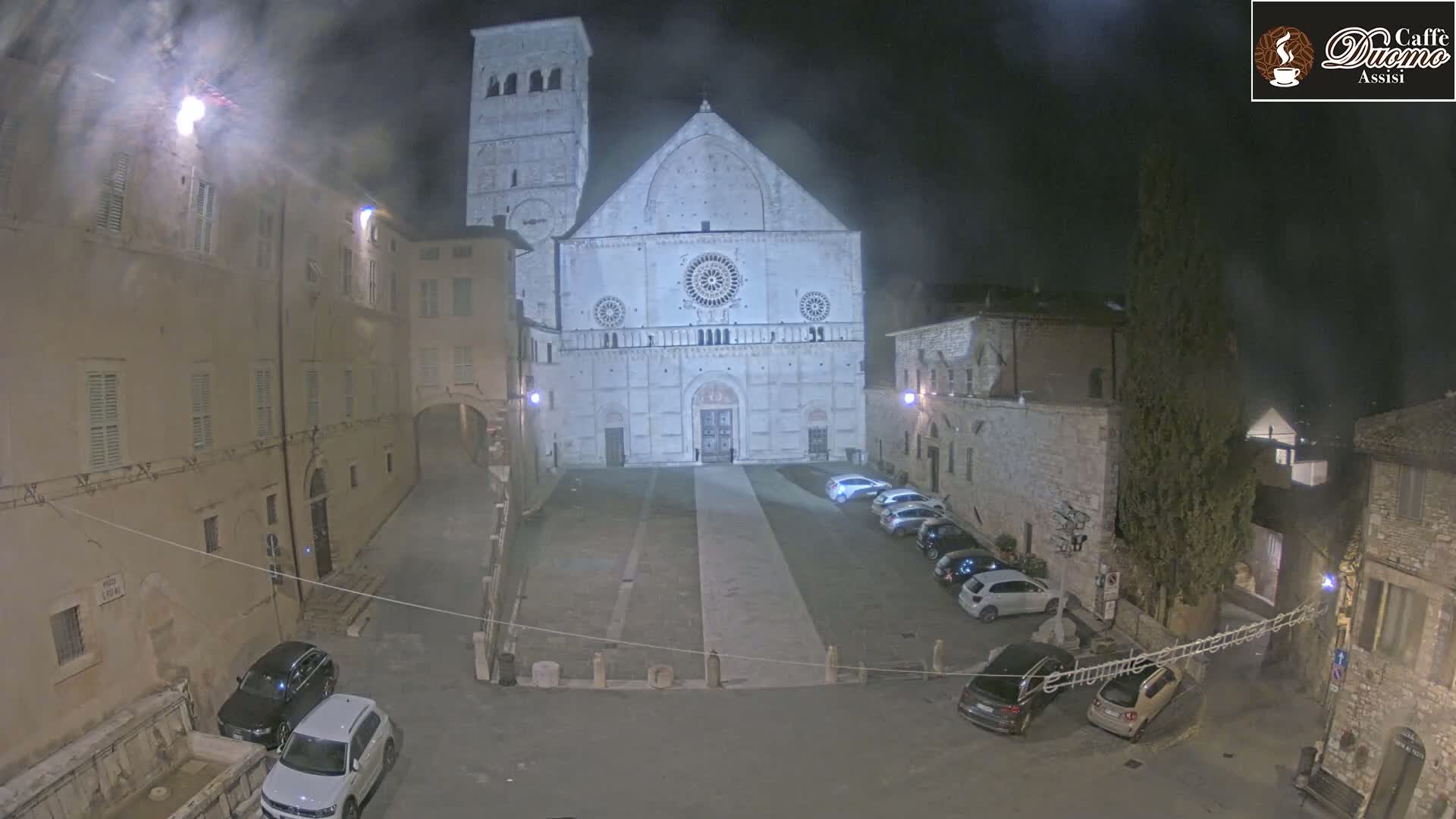 At night, under hazy conditions, a large stone cathedral stands in a town square with several cars parked nearby.