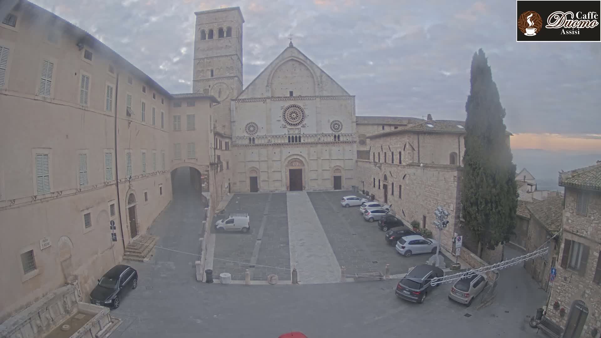 A stone plaza with several parked cars sits in front of a large cathedral, under an overcast sky.