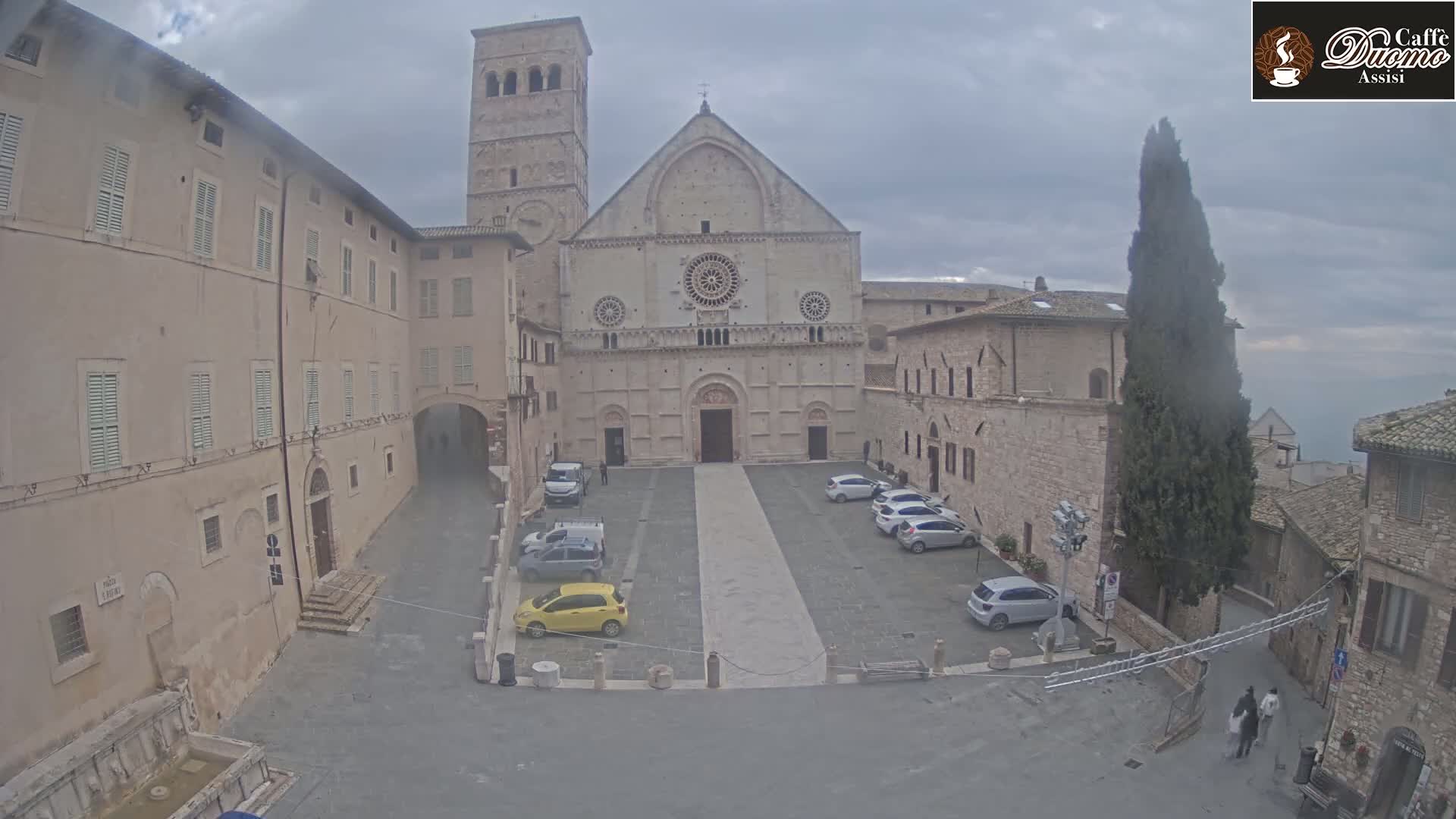 A large stone church with a tall bell tower sits in a courtyard with several parked cars, under an overcast sky.