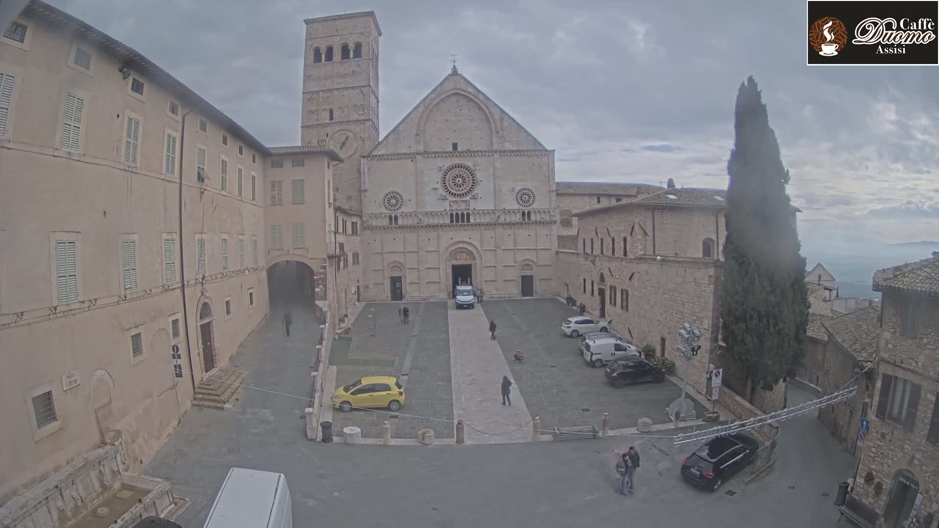 A large stone cathedral stands in a plaza surrounded by buildings, under an overcast sky.