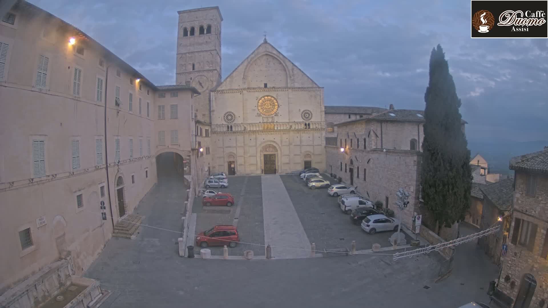 A large stone cathedral sits in a town square with several cars parked, under a cloudy, dusky sky.