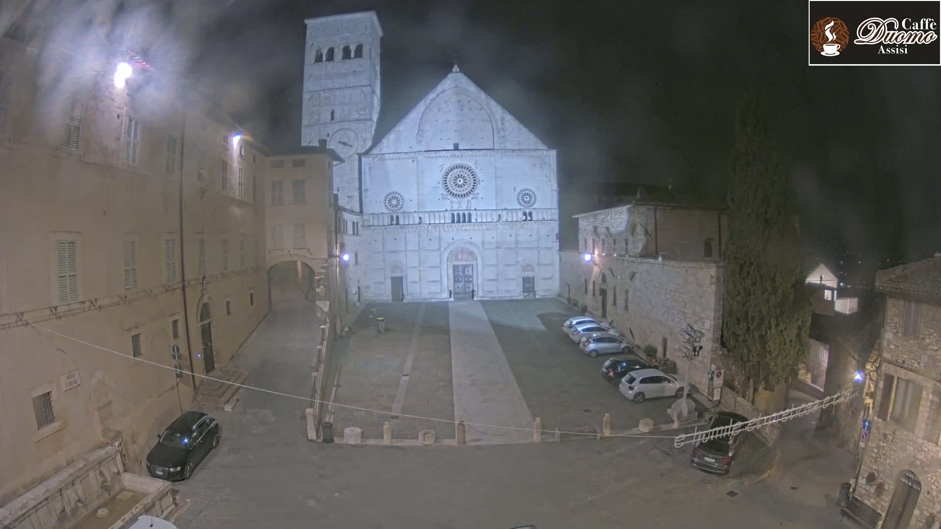 A large cathedral is illuminated at night in a town square with several parked cars under overcast skies.