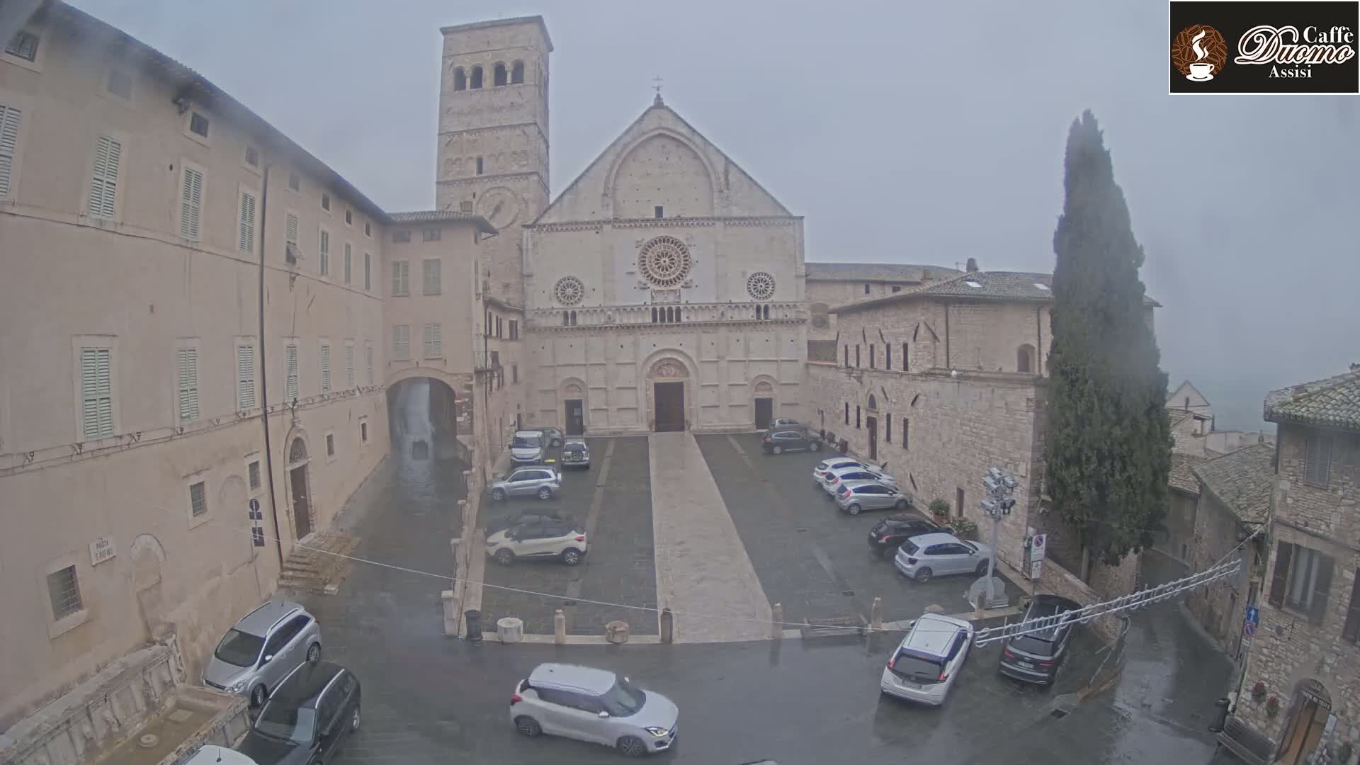A large stone cathedral sits in a town square with several cars parked, all under a gray, rainy sky.