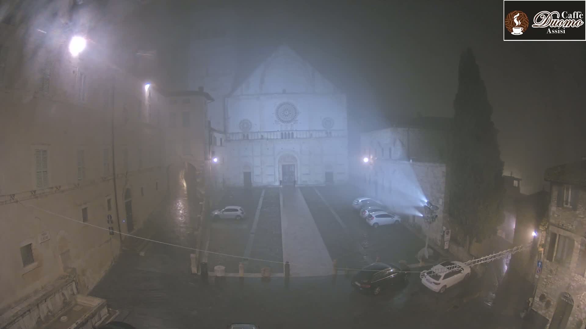 A large stone church is visible through heavy fog and rain at night, surrounded by parked cars and buildings in a town square.