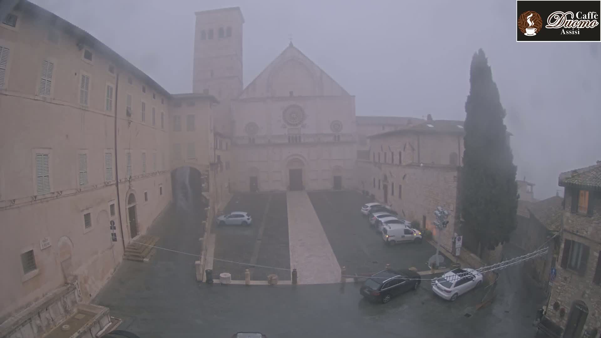 A large, stone church is visible in a town square on a foggy, rainy day, with cars parked nearby and tall buildings surrounding the area.