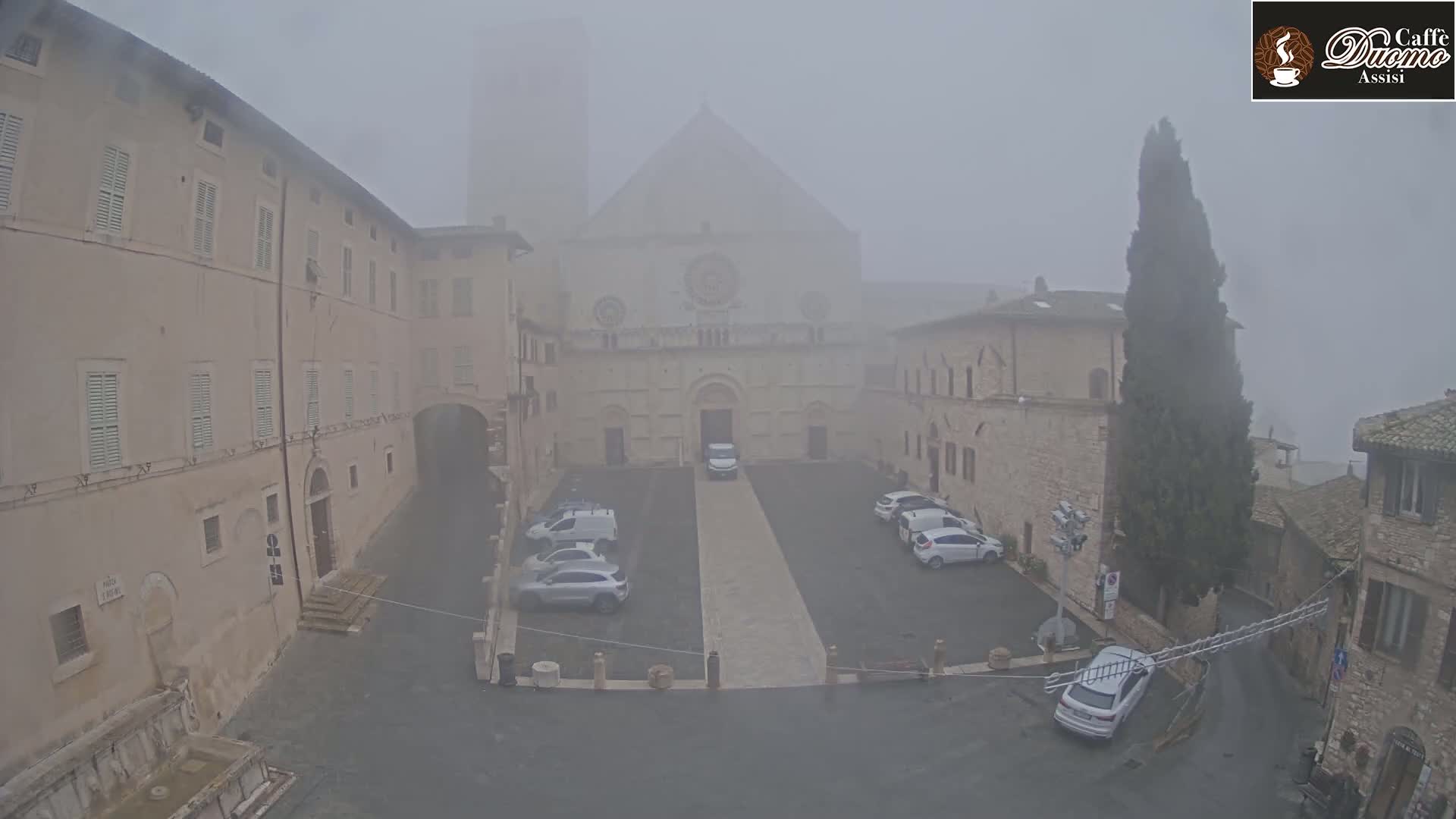 A foggy view of a square with parked cars, surrounded by stone buildings, and a church in the background.