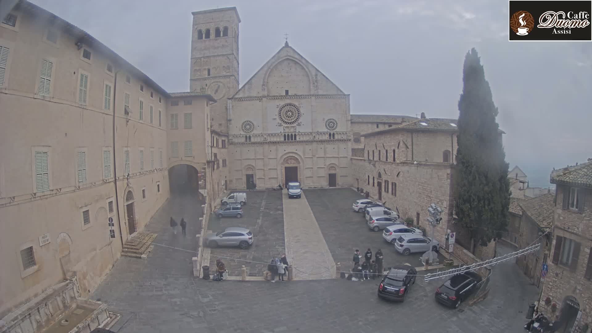 A large stone church is situated in a town square with several cars parked, under an overcast sky.