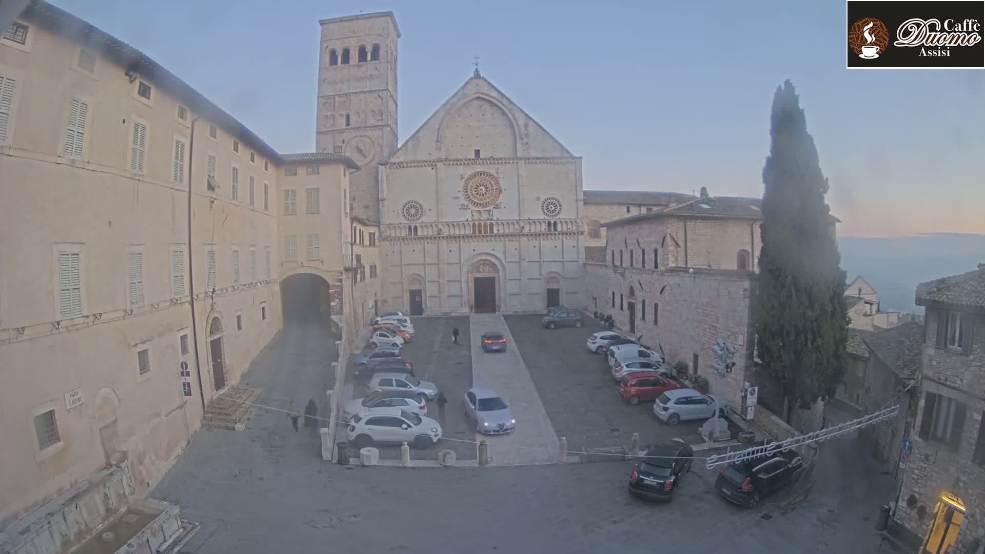 A large stone cathedral sits in a town square with several cars parked, under a hazy, dusky sky.