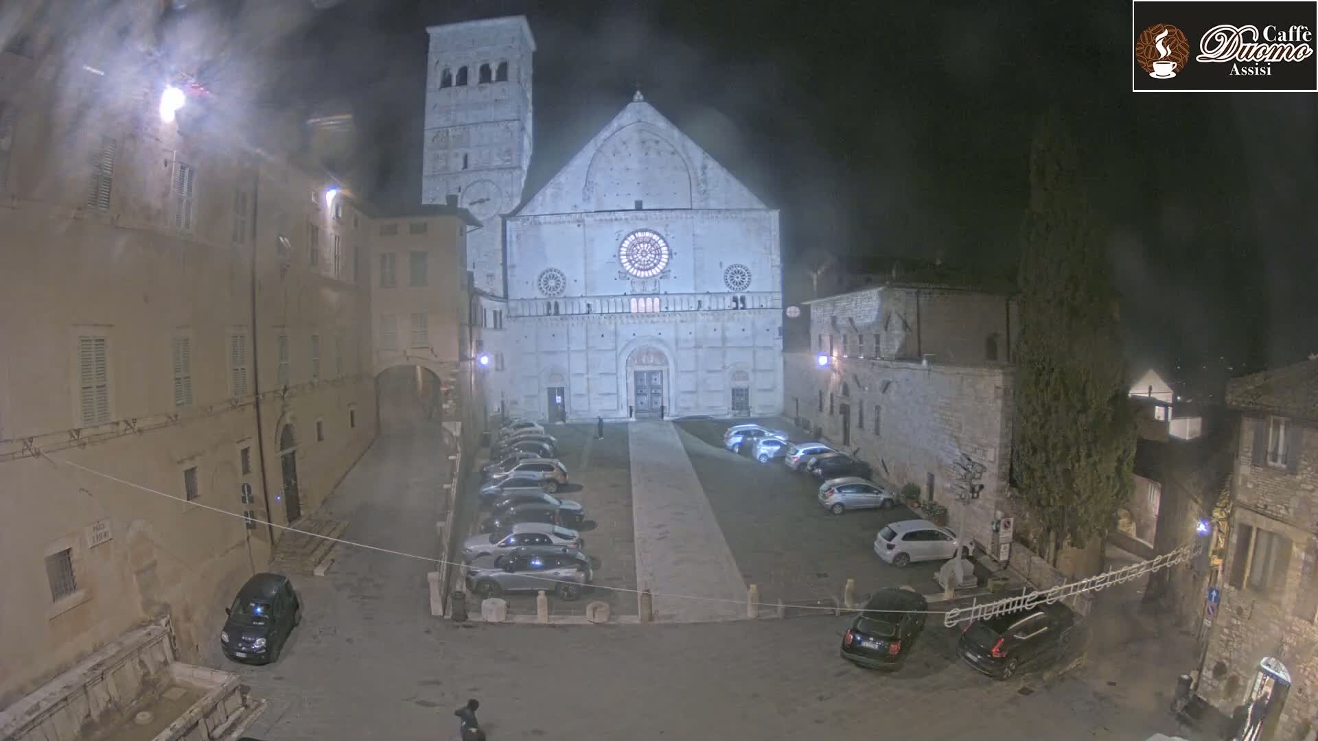 A nighttime view of a cathedral and surrounding square with cars parked, under a hazy sky.