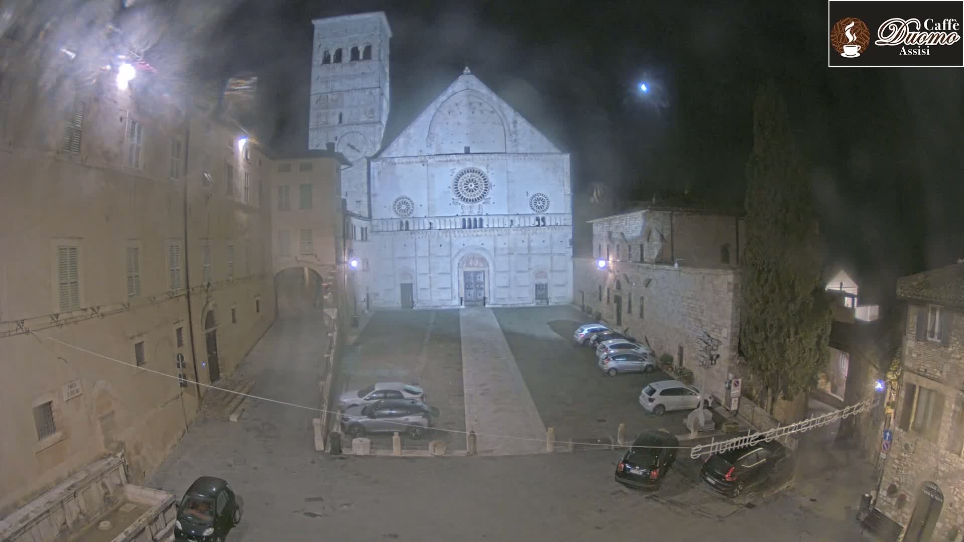 At night, under a hazy sky, a large stone cathedral stands in a town square with several cars parked in front of it and older buildings surrounding the area.