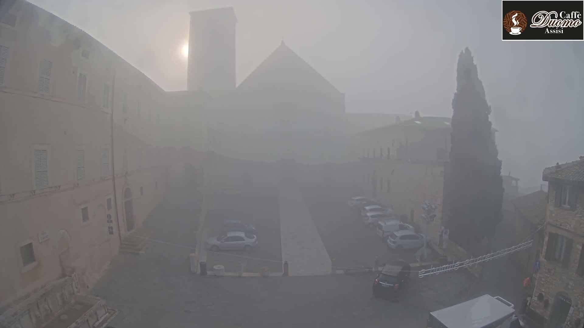 A foggy square in a town, showing several cars parked and tall buildings in the background, with the sun barely visible through the mist.