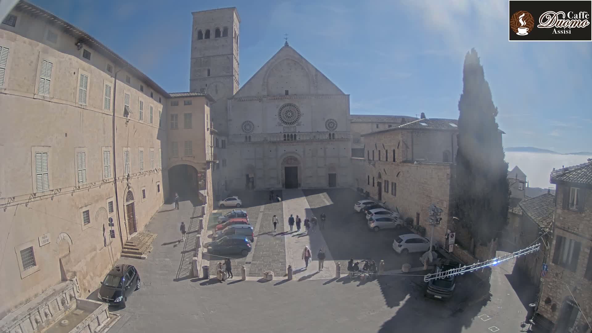 A large stone cathedral stands in a sunlit town square surrounded by older buildings, with a layer of fog visible in the distance.