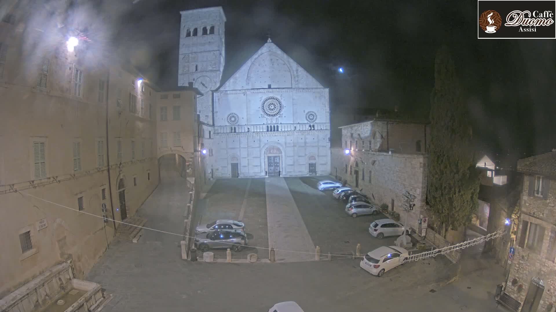 At night, a large stone cathedral stands in a plaza with several cars parked, and older stone buildings surround it under a hazy, dark sky.