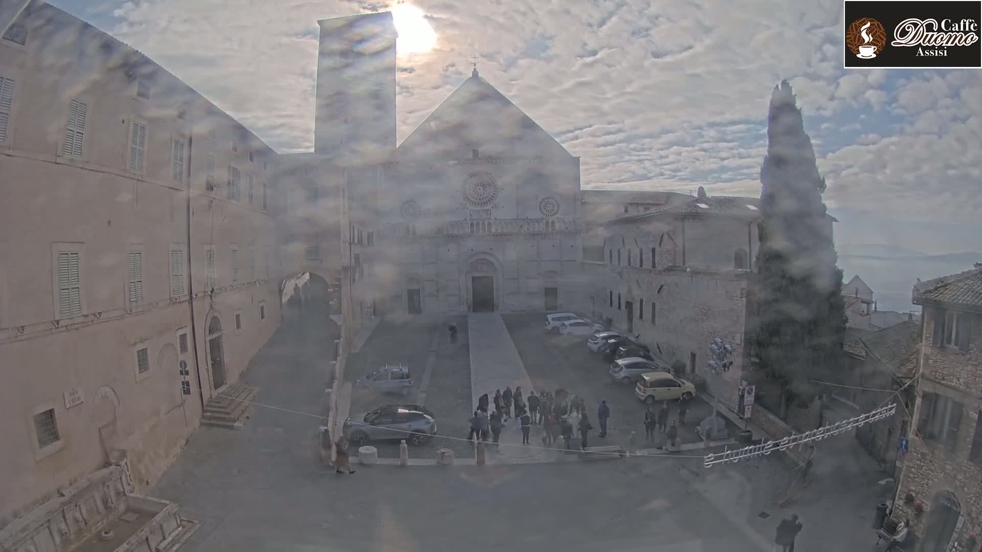 A hazy sun shines on a large stone cathedral in a town square, surrounded by buildings and parked cars, on a cloudy day.