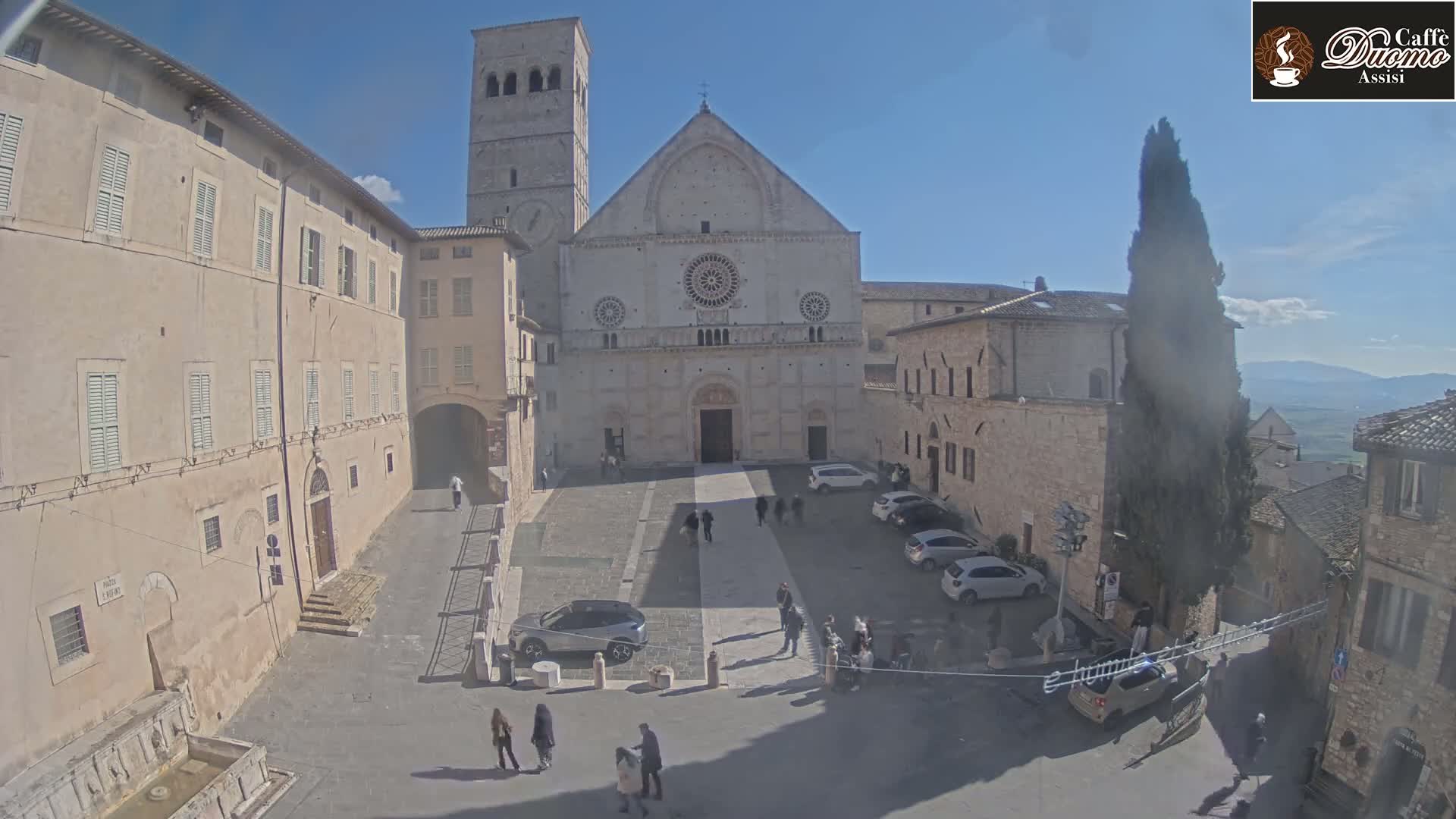 A sunny day reveals a large stone plaza, centered on a cathedral with a tall bell tower, surrounded by old stone buildings and a few parked cars.