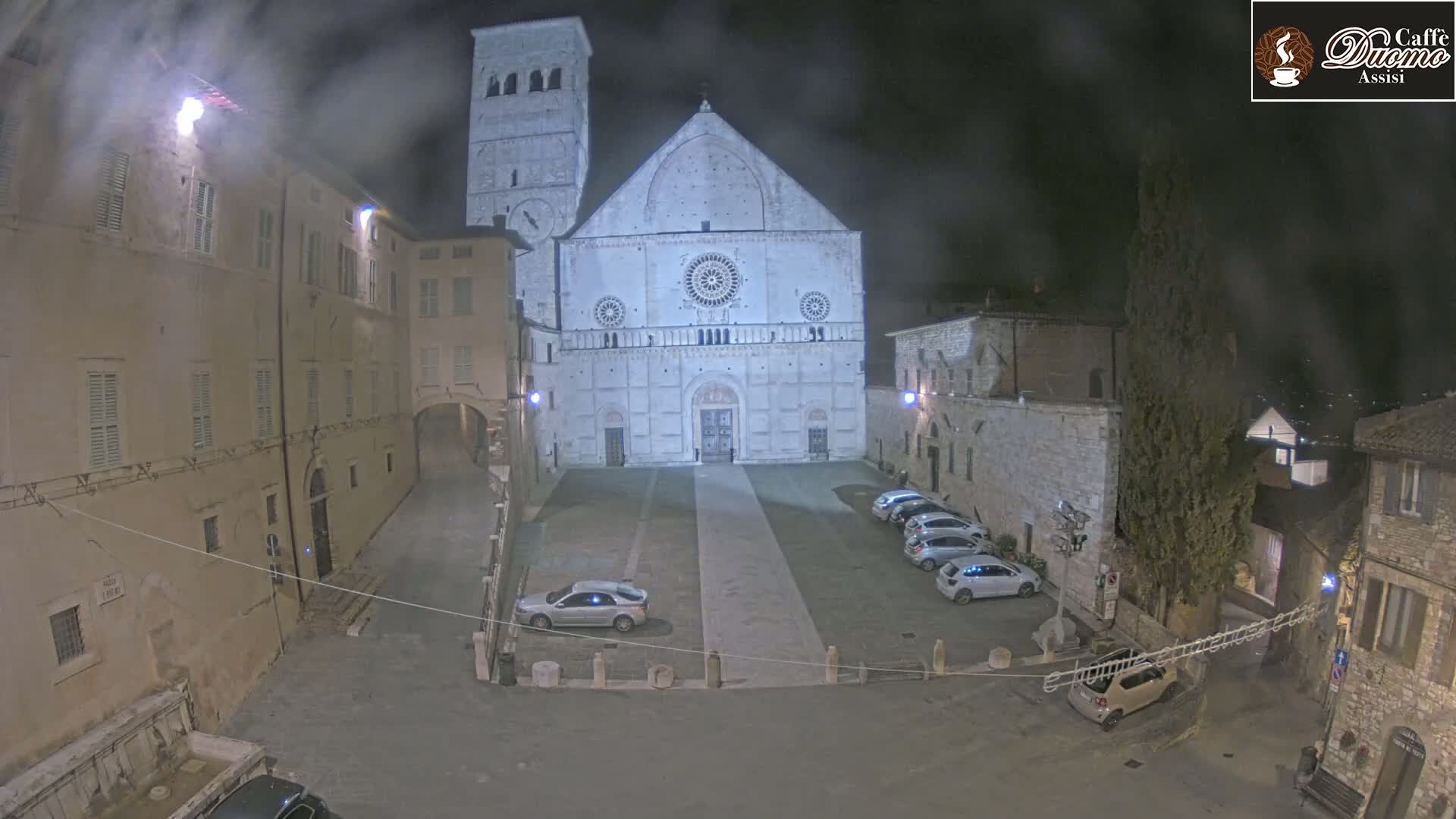 A large, light-colored cathedral stands in a plaza at night, surrounded by older stone buildings, under hazy or foggy conditions.