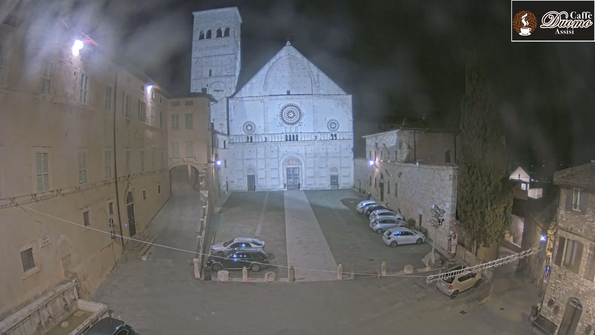 A large, light-colored cathedral stands in a dimly lit plaza at night, surrounded by buildings and several parked cars under a hazy sky.