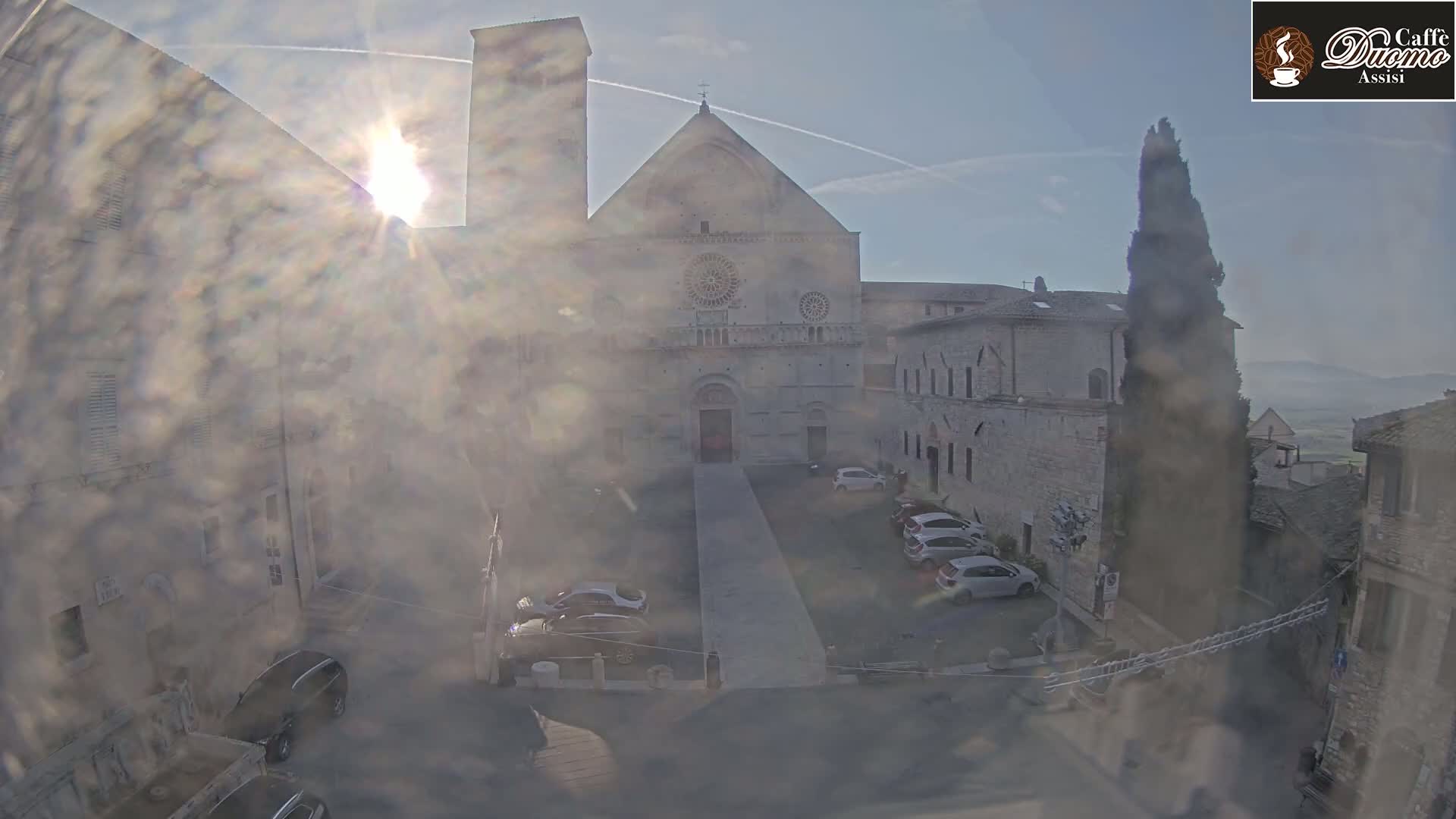 A hazy, sunny view of a stone cathedral and surrounding buildings with a few cars parked in a courtyard.