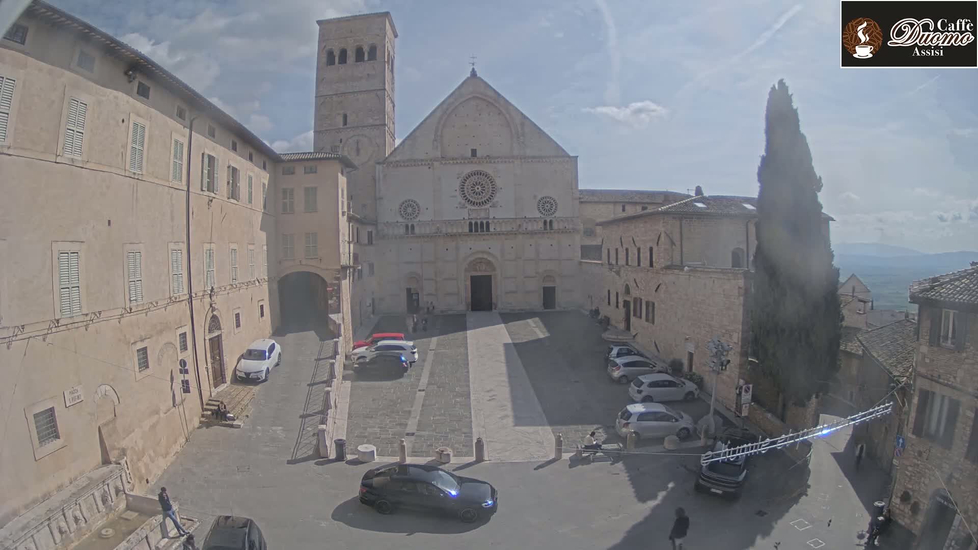 A town square with a large cathedral in the background, under a partly cloudy sky, contains several parked cars and a few pedestrians.