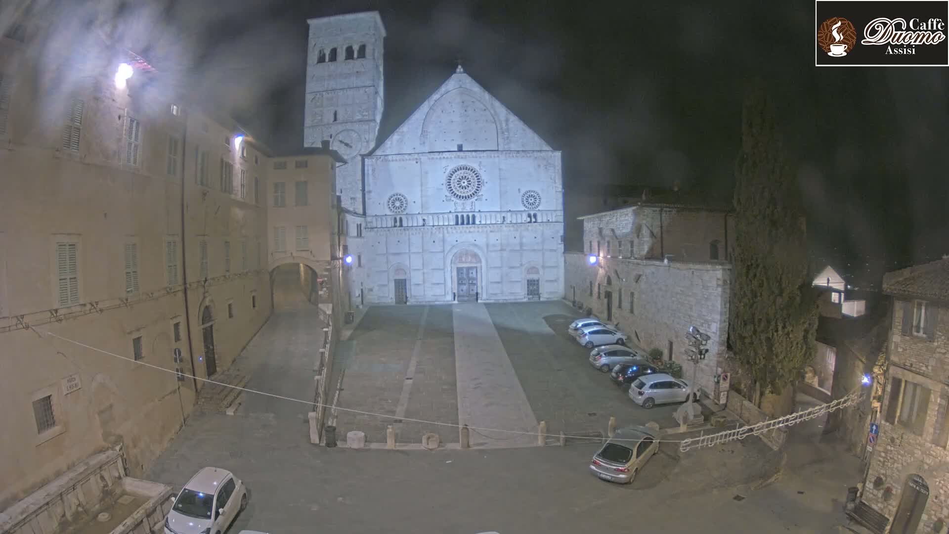 A nighttime, overcast view of a large stone cathedral in a town square with several cars parked, surrounded by older stone buildings.