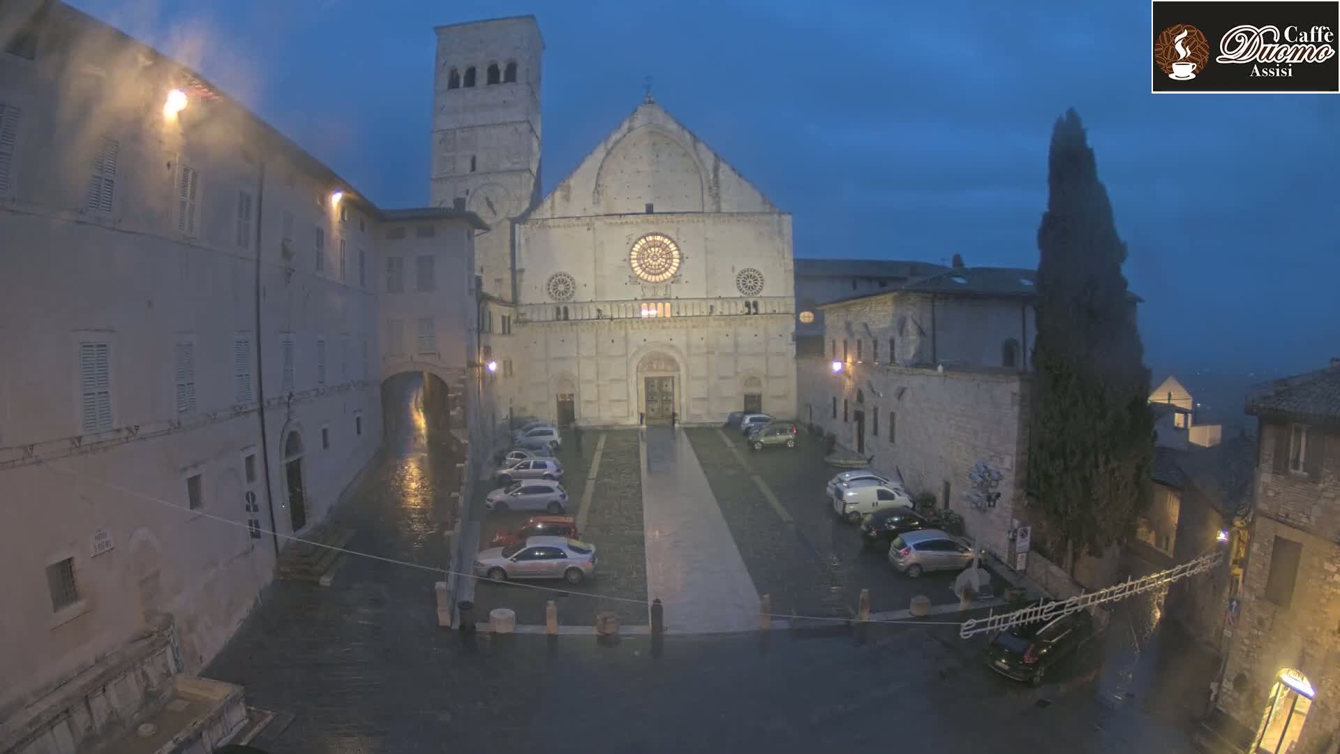 A large stone cathedral is situated in a town square at night during a light rain, with cars parked in the square and buildings lining the perimeter.