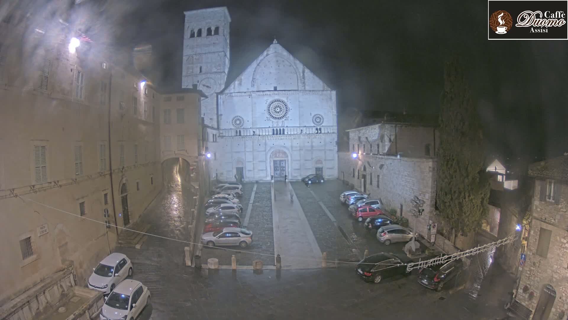 A large, light-colored cathedral is situated in a town square at night under rainy conditions, with cars parked on either side of a pathway leading to its entrance.
