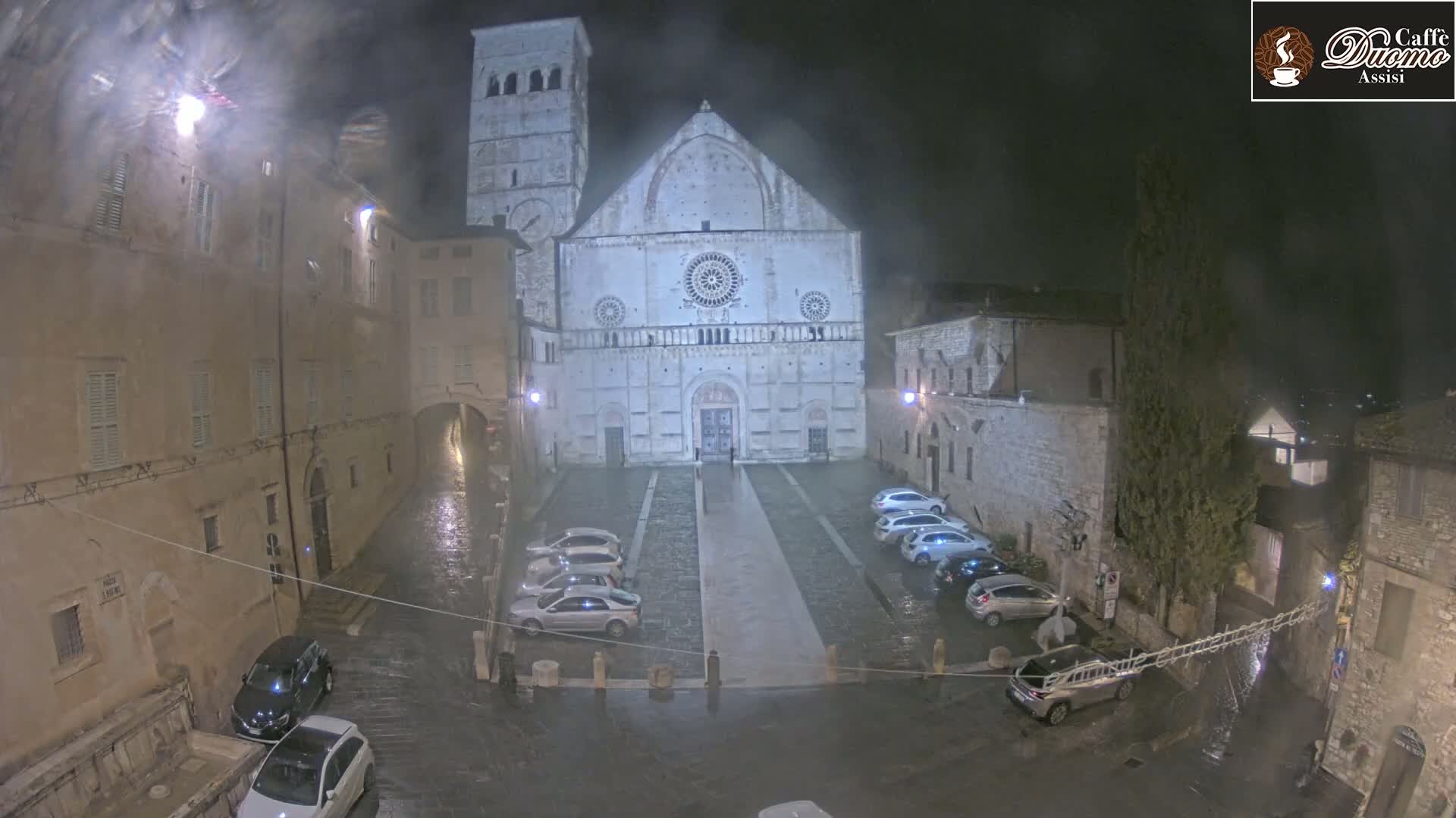 A large stone cathedral stands in a town square at night during a rainstorm, surrounded by parked cars and older buildings.