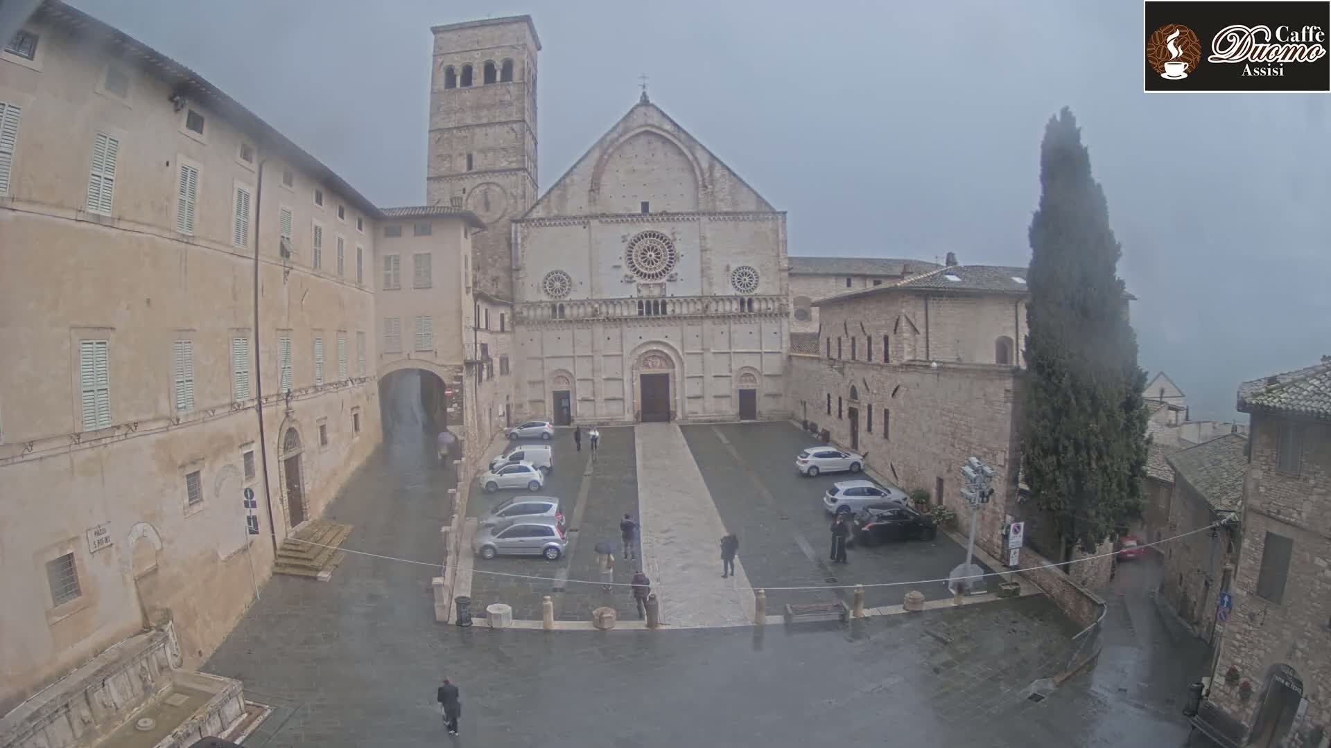 A large stone church, surrounded by buildings, sits in a wet square where several cars are parked under an overcast sky.