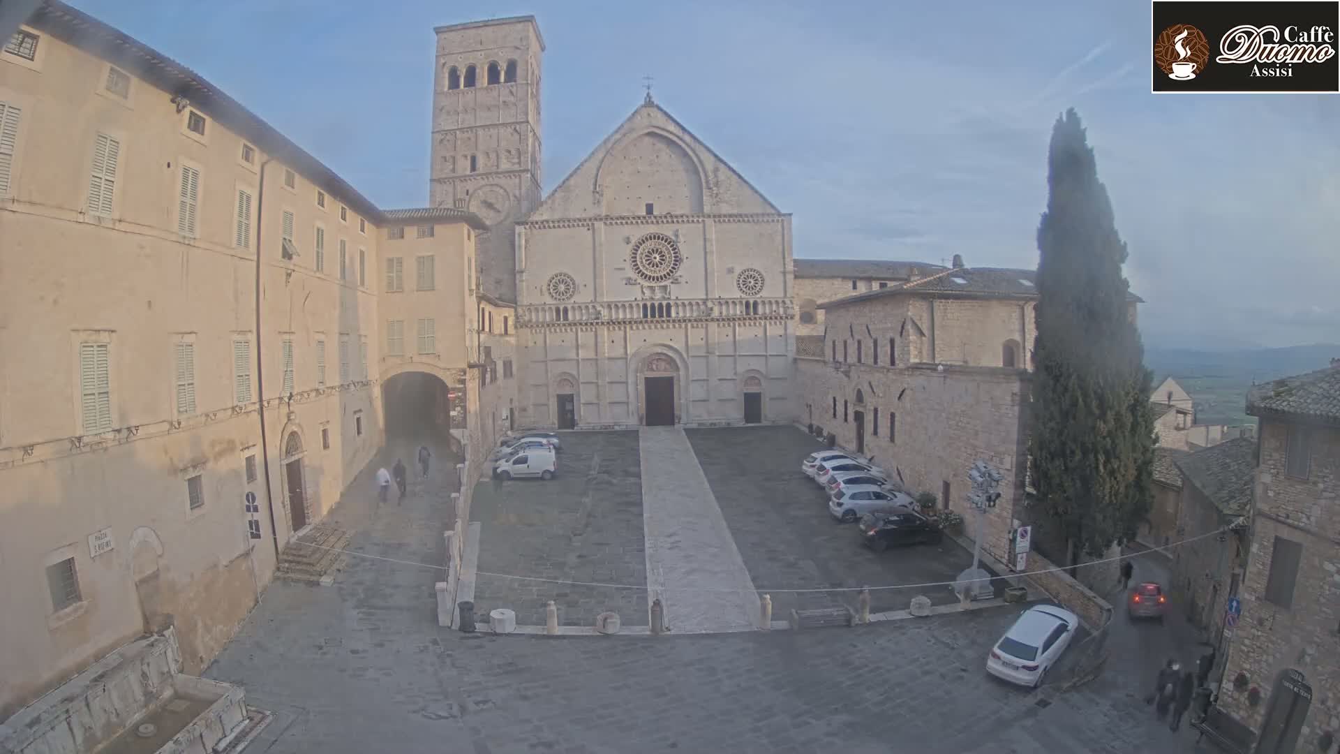 A stone plaza with parked cars, surrounded by aged buildings, including a large cathedral, under a mostly clear sky.