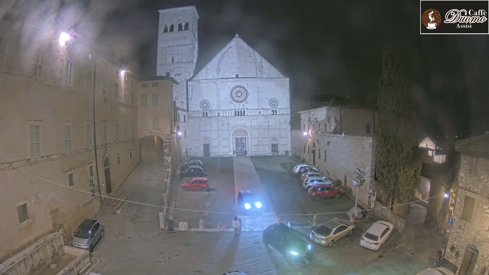 A nighttime, slightly hazy view of a town square with a large cathedral, several parked cars, and surrounding buildings.