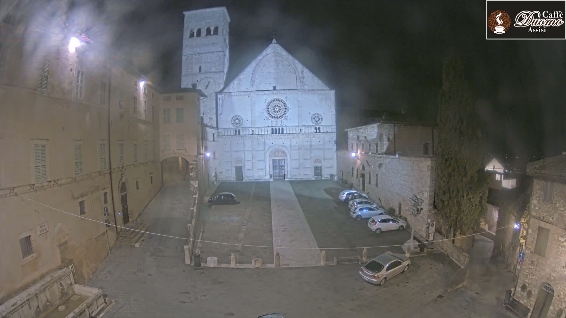 A nighttime view of a town square with a large cathedral at its center, under a misty or hazy sky.