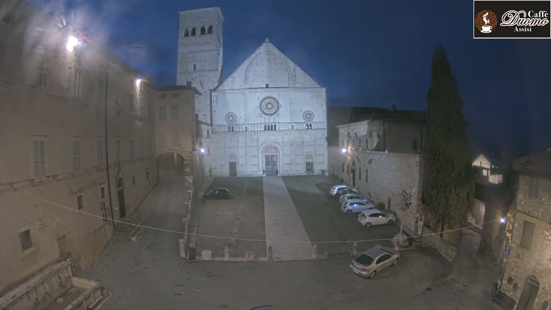 A large, light-colored cathedral stands in a stone plaza at night, with several cars parked nearby and buildings lining the square under a dark, clear sky.
