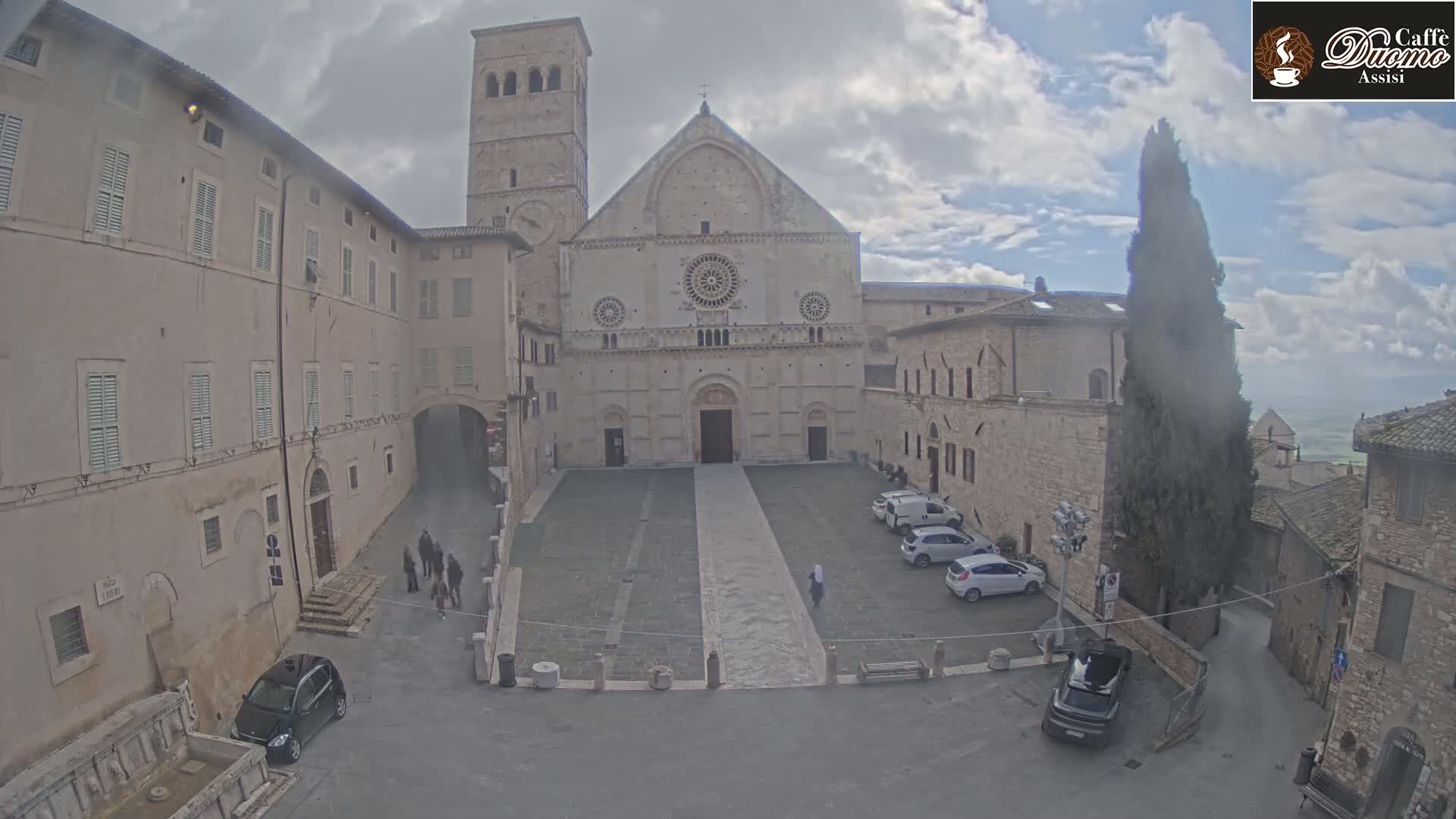 A large stone cathedral stands in a town square under a cloudy sky, with several cars parked nearby and a few pedestrians walking around.