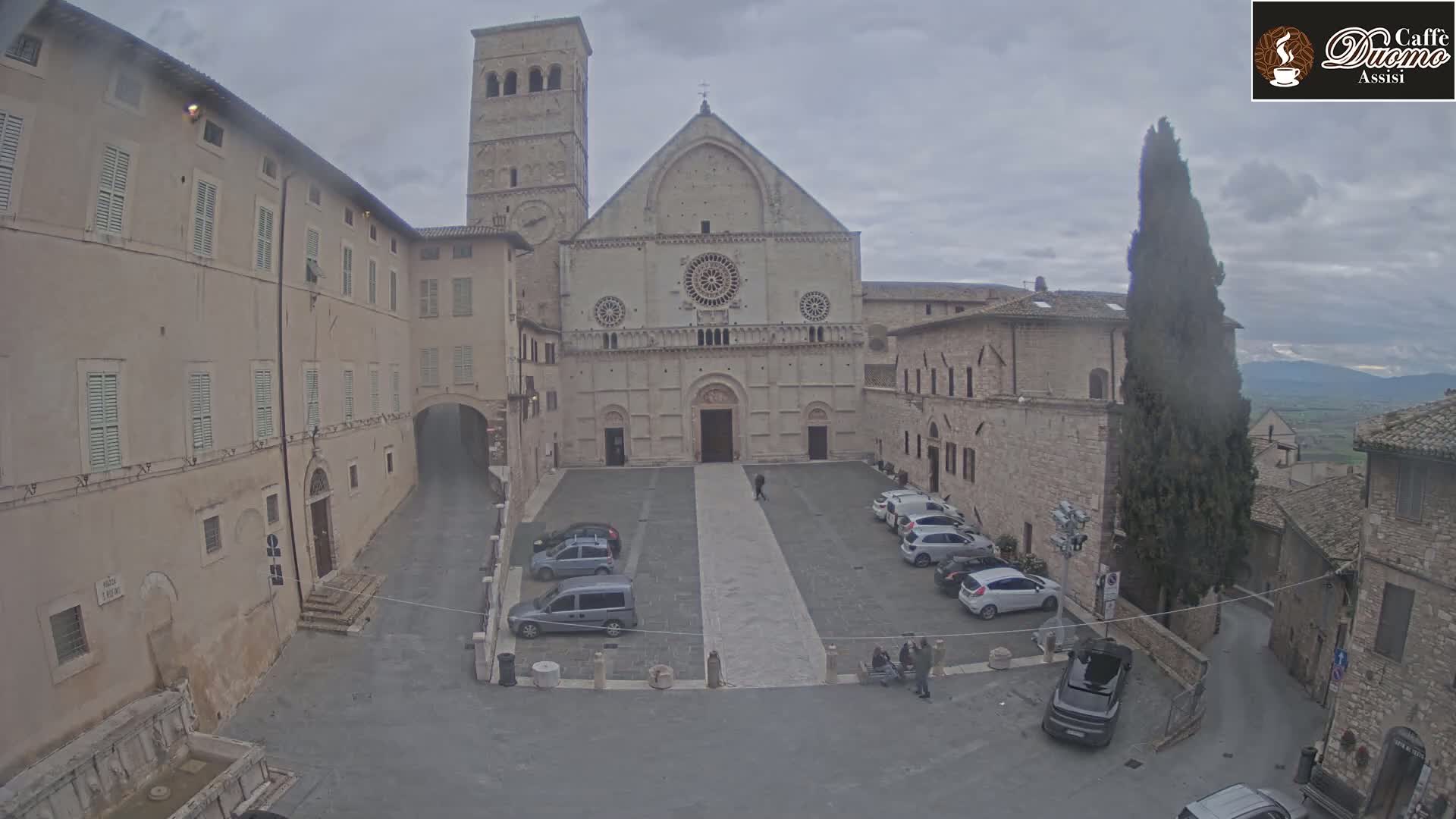 A large stone cathedral sits in a town square under an overcast sky, surrounded by other stone buildings and a few parked cars.