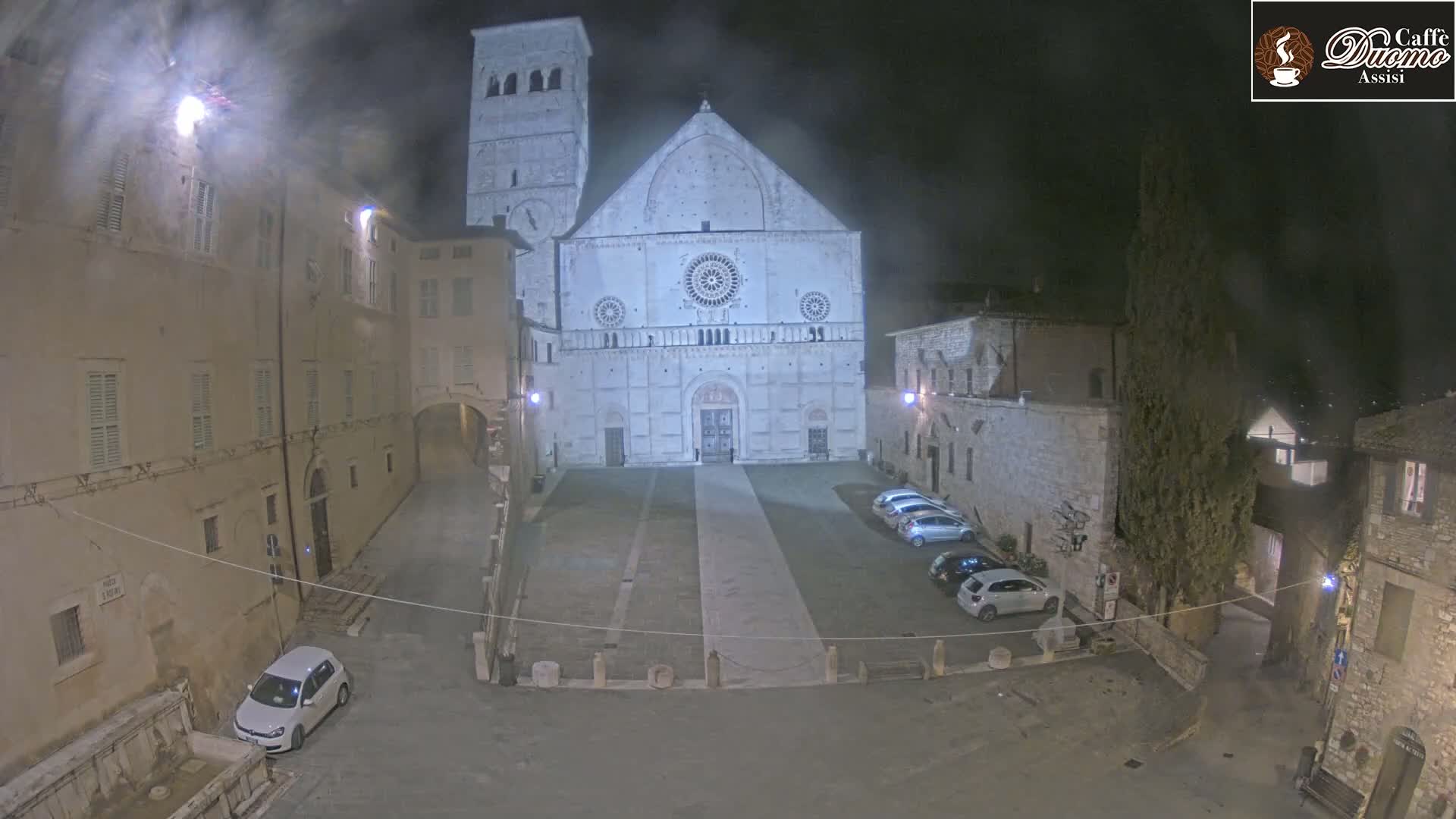 A large, light-colored cathedral stands in a stone plaza at night, several cars parked nearby, under a cloudy sky.