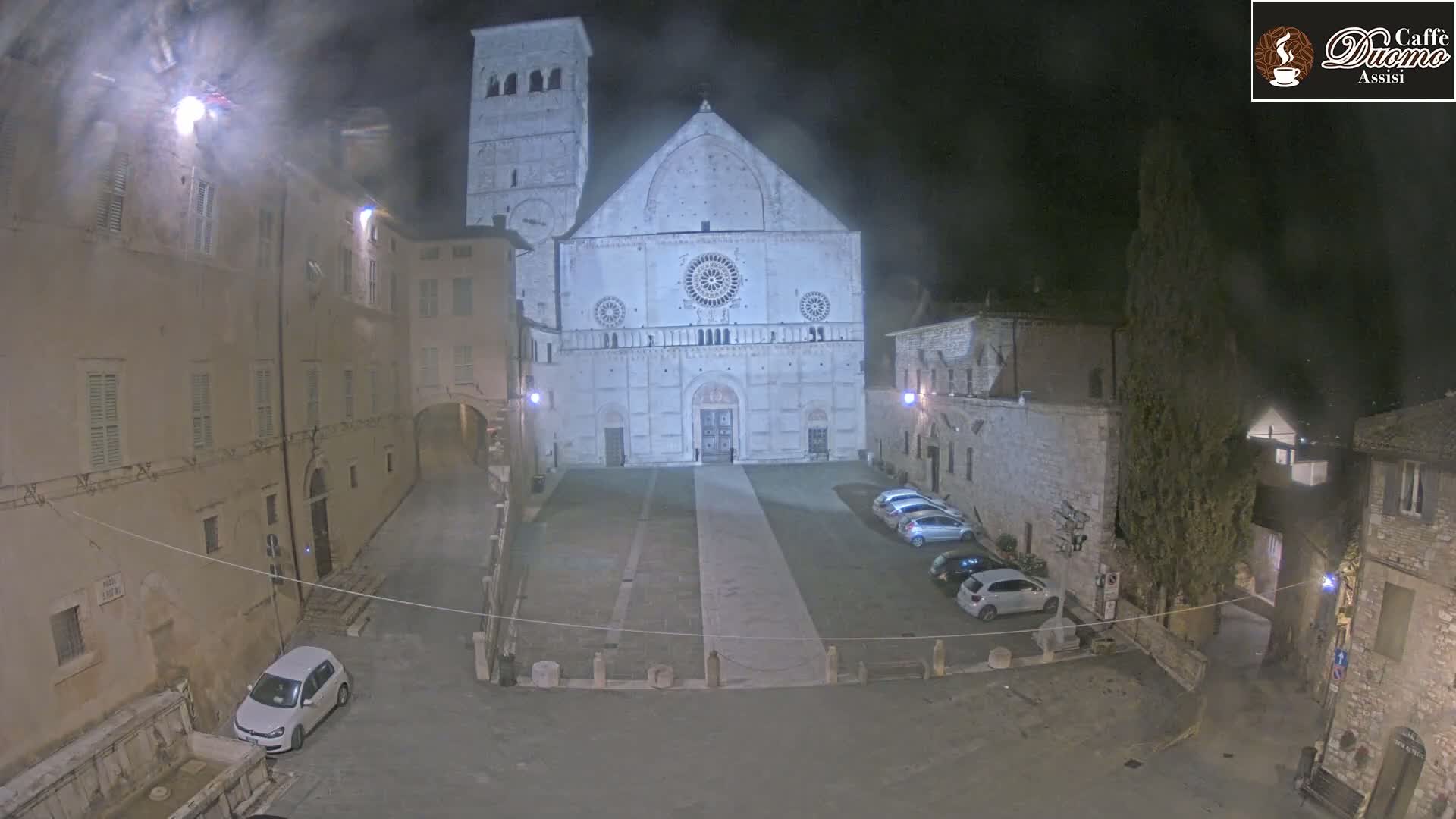 A large stone cathedral is seen at night in a dimly lit plaza with several cars parked nearby, under hazy conditions.